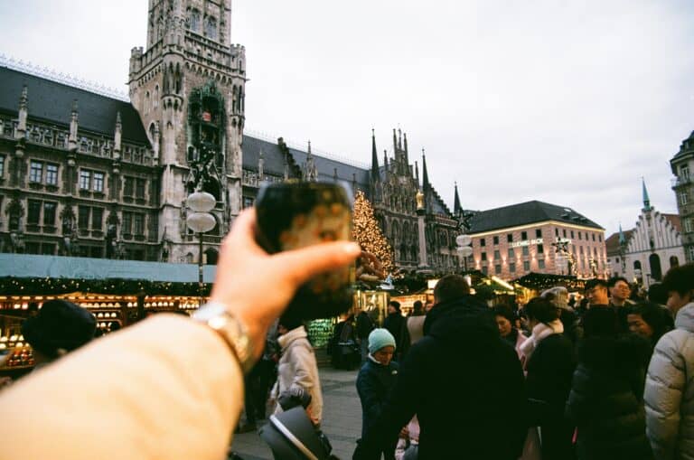 Eine Tasse Glühwein wird von einer Frau auf einem Weihnachtsmarkt Hochgehalten im Hintergrund sieht man das Rathaus von München